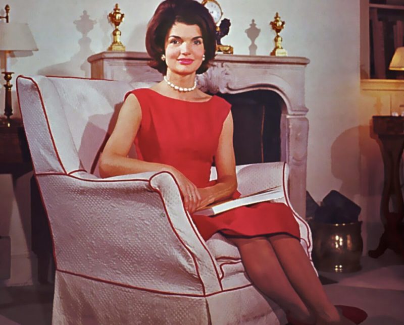 a woman sitting in a white chair wears a red dress and a pearl necklace